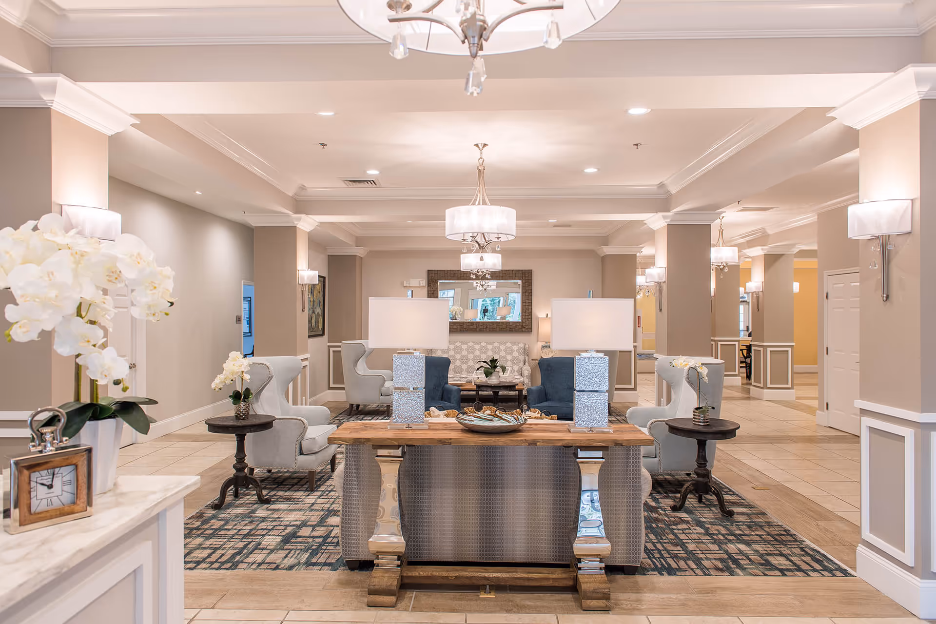 A spacious and elegantly decorated senior living facility common area with beige walls, tiled floors, and multiple seating arrangements including armchairs and sofas. The room features modern chandeliers, table lamps, a large mirror on the far wall, and decorative plants and flowers on tables.