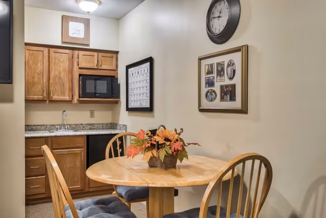 A small dining area with a round wooden table and two wooden chairs with blue cushions. On the table is a basket with artificial autumn leaves and flowers. Behind the table is a kitchenette with wooden cabinets, a granite countertop, a sink, a microwave, and a small refrigerator. On the wall above the table are a large round clock and a framed collage of photos. There is also a framed calendar on the wall near the kitchenette.