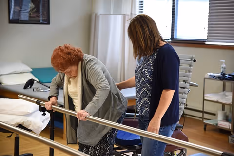 An elderly woman using parallel bars for physical therapy assistance while a caregiver supports her from behind in a therapy room with medical equipment and a wheelchair nearby.