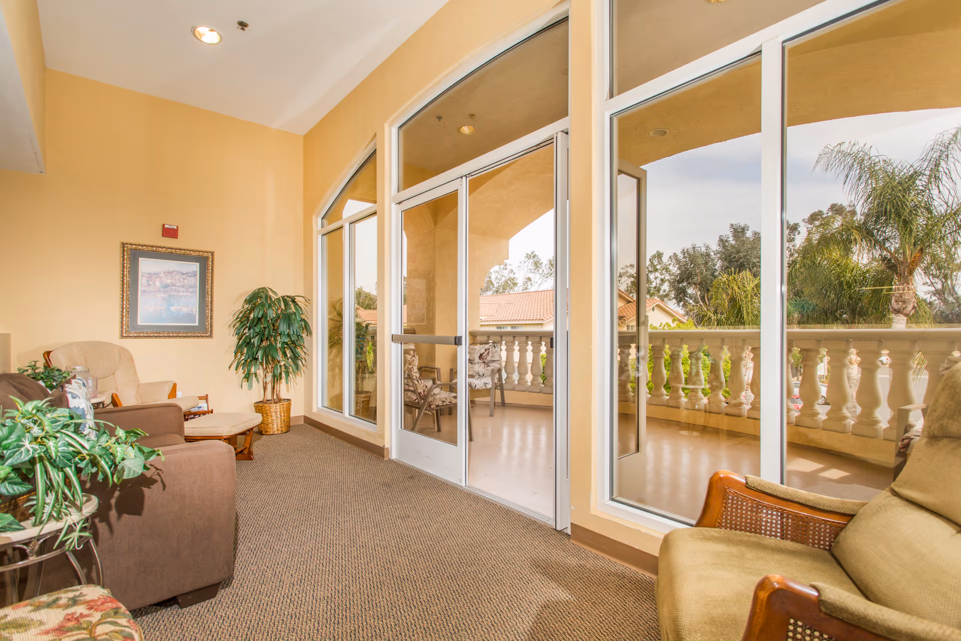 Sunlit seating area with armchairs, plants and large glass doors opening onto a balcony with a balustrade.