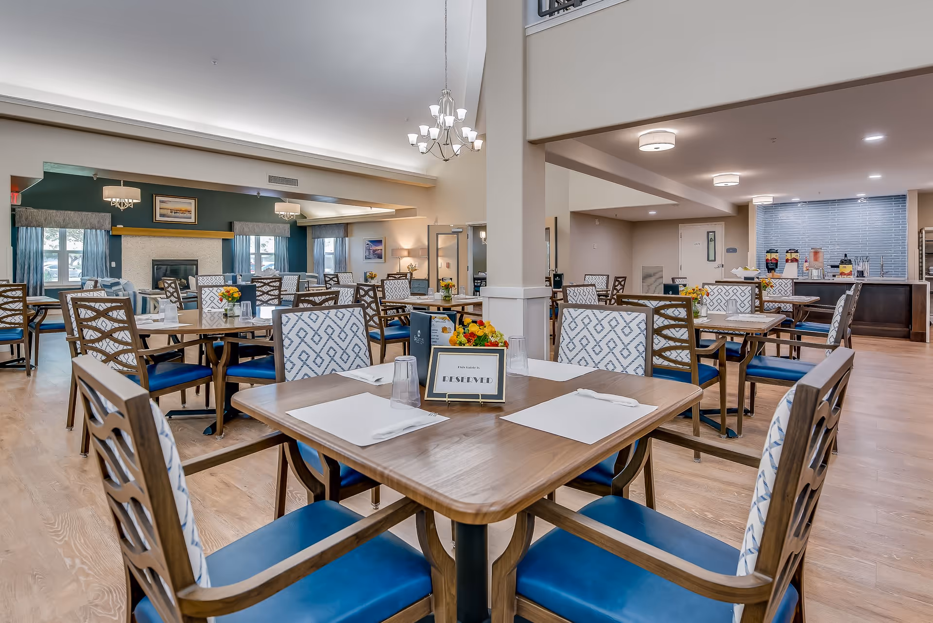 A spacious and well-lit dining room in a senior living facility with multiple wooden tables and chairs featuring blue cushions and patterned backs. Each table is set with placemats, napkins, and glasses, and some have small flower arrangements and reserved signs. The room has wooden flooring, a fireplace with framed artwork above it, large windows with curtains, and a buffet area with beverage dispensers in the background.