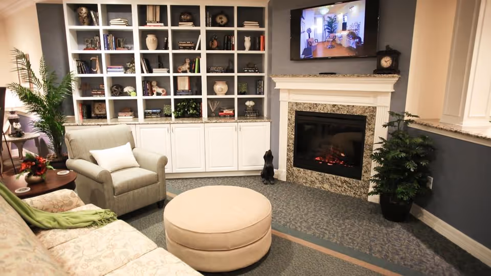 Cozy living room with armchairs, a round ottoman, built-in shelves, and a fireplace with a TV mounted above.