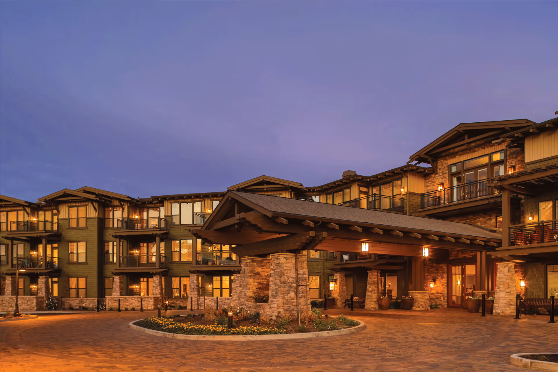 Exterior view of Fountaingrove Lodge at dusk, showing a large multi-story building with warm lighting, stone pillars, balconies, and a covered entrance with a circular driveway and landscaped flower beds.