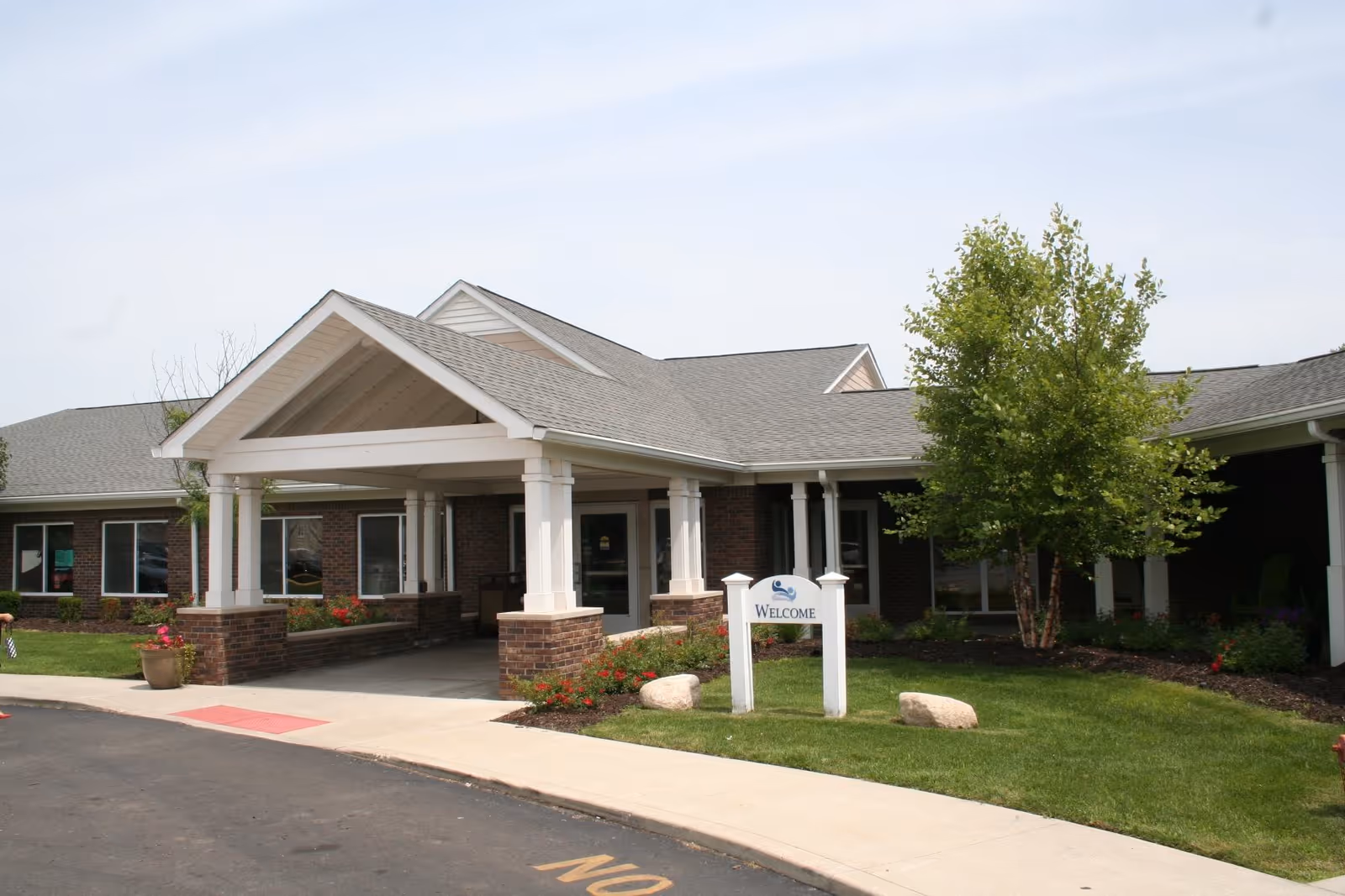 Exterior view of a single-story brick building with a covered entrance supported by white columns. There is a small landscaped area with green grass, a tree, and flowers near the entrance. A white sign with the word 'WELCOME' is placed on the lawn. The sky is clear with some light clouds.