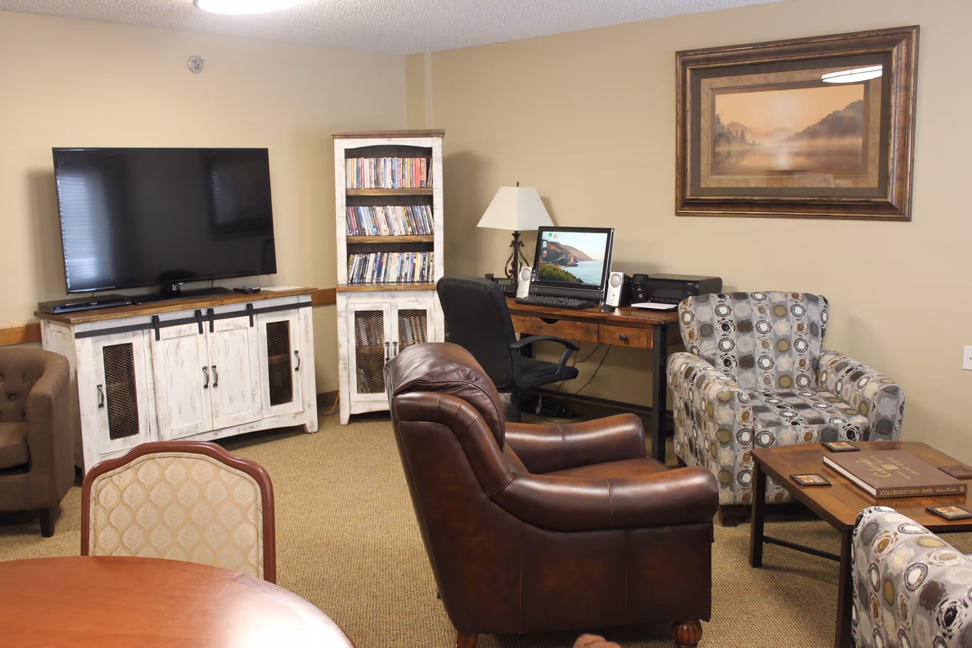 A cozy living room area with a large flat-screen TV on a white distressed cabinet, a tall white distressed bookshelf filled with DVDs, a wooden desk with a computer and lamp, a brown leather armchair, a patterned armchair, a wooden coffee table with books, and a framed landscape painting on the beige wall.