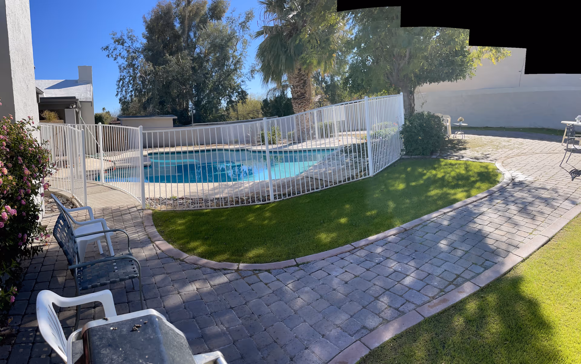 Outdoor area with a fenced swimming pool surrounded by a paved walkway and green grass. There are several chairs and a bench along the walkway, with trees and bushes providing shade and greenery.