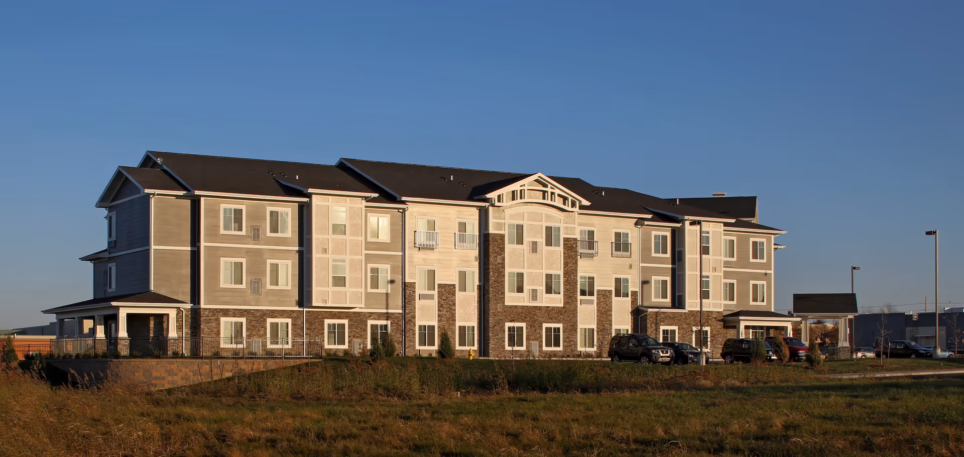 Exterior view of a three-story senior living facility building with multiple windows, a combination of stone and light-colored siding, and a dark roof under a clear blue sky. Several cars are parked in front of the building, and there is a grassy area in the foreground.