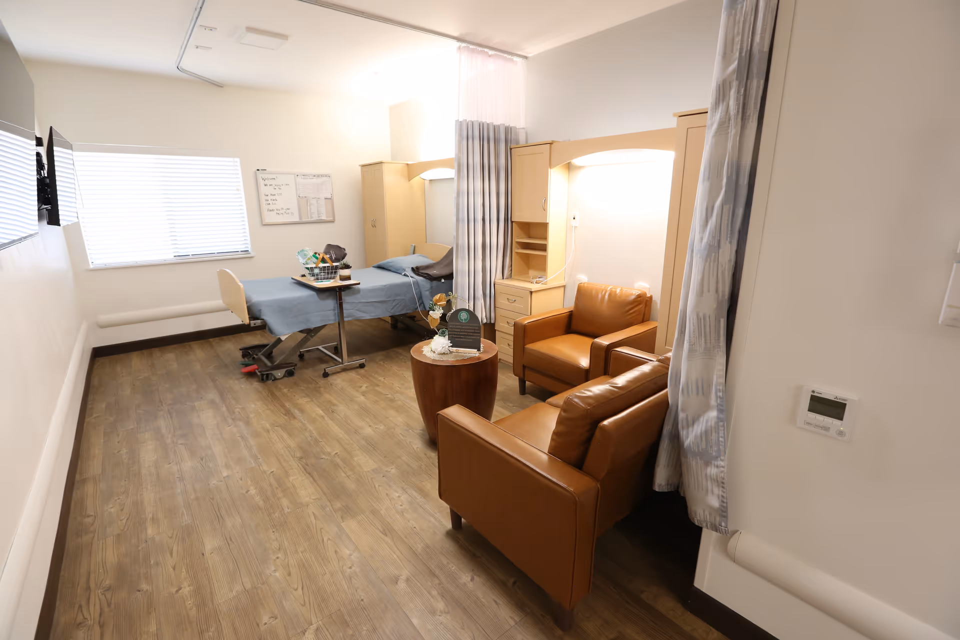 A well-lit room in a senior living facility featuring a hospital-style bed with blue bedding, a small table with a basket and flowers, two brown leather armchairs, a wooden side table with a decorative item, a window with blinds, and a privacy curtain partially drawn between the bed and seating area.