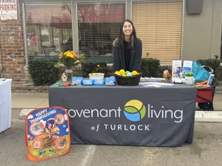 A person stands behind a table draped with a Covenant Living of Turlock tablecloth displaying brochures, fruit, and fall decorations outside a building.