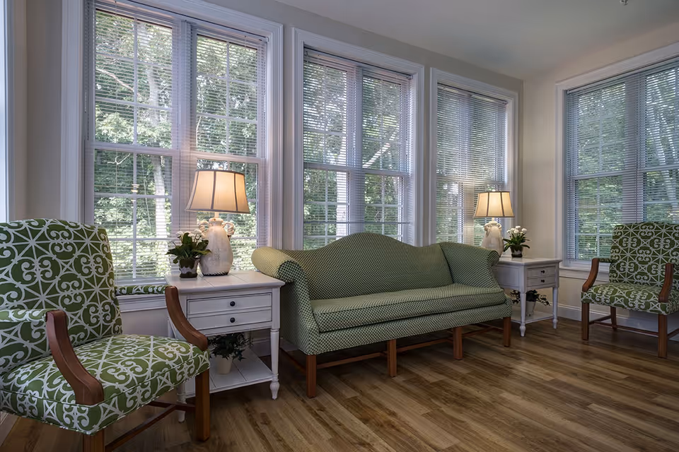 Sunlit sitting area with a green upholstered sofa and patterned chairs flanking white side tables and lamps in front of large windows.