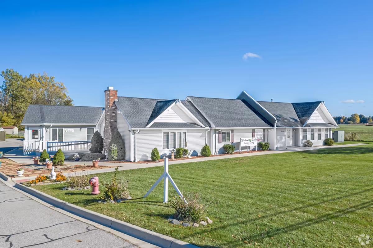 Single-story white senior living building with pitched roofs, a landscaped front lawn, and driveway under a clear blue sky.