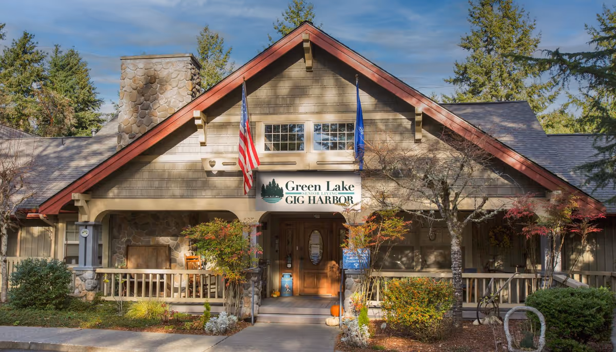 Front exterior view of Greenlake Senior Living Gig Harbor building with a peaked roof, stone chimney, American and state flags, a wooden porch with chairs, and surrounding trees and shrubs.