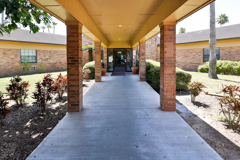 Covered walkway with brick pillars leading to the entrance of a single-story brick building surrounded by landscaped bushes and palm trees under a clear sky.