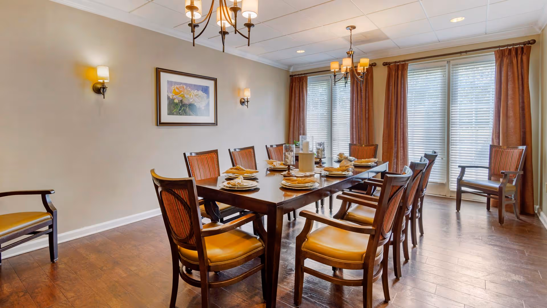 A dining room with a long wooden table set for a meal with plates, napkins, and glasses. The room has wooden chairs with yellow cushions around the table and additional chairs along the walls. There are large windows with blinds and brown curtains, wall sconces, and a chandelier providing warm lighting. A framed floral painting hangs on the beige wall.