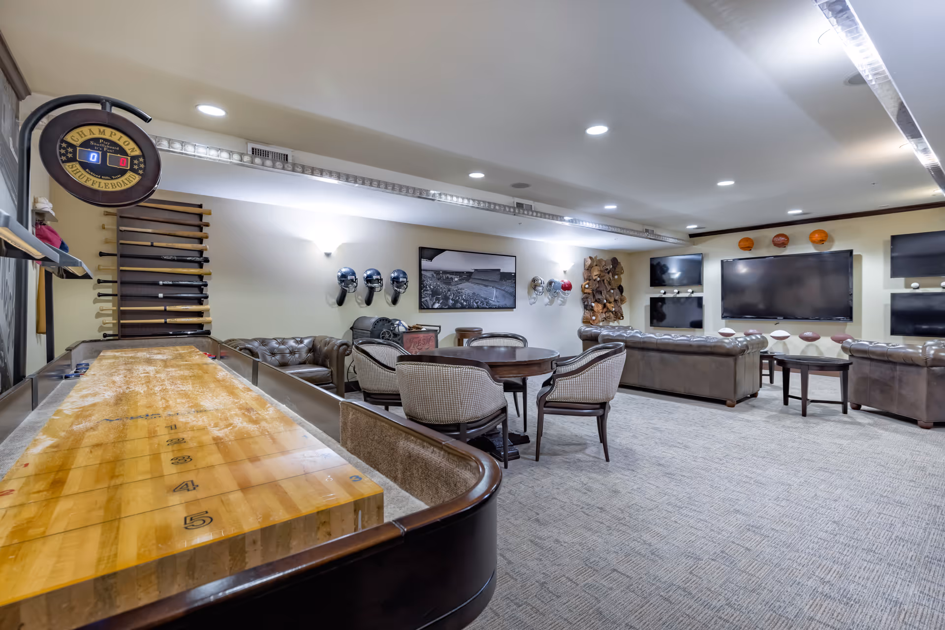 A spacious recreational room with a shuffleboard table in the foreground, several comfortable chairs and sofas arranged around a round table and multiple large flat-screen TVs mounted on the walls. Sports memorabilia, including helmets, basketballs, and footballs, decorate the walls and shelves.