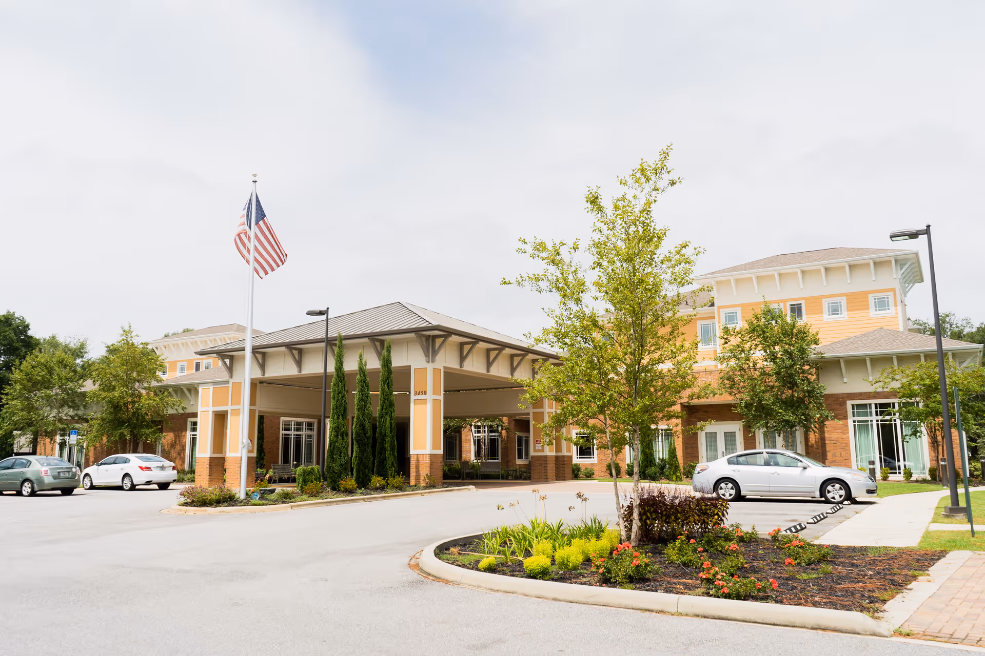 Front entrance of a two-story assisted living building with a covered porte-cochère, American flag, parked cars, and landscaped grounds.
