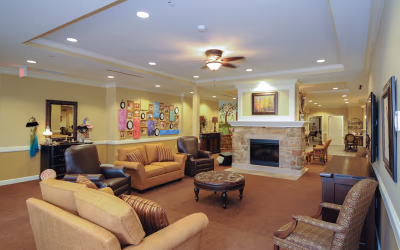 A cozy living room area in a senior living facility with multiple upholstered chairs and sofas arranged around a central round ottoman. There is a stone fireplace with a framed painting above it, warm lighting from ceiling fixtures and lamps, and decorative artwork on the walls. The space has a warm color palette with beige walls and brown carpeting.