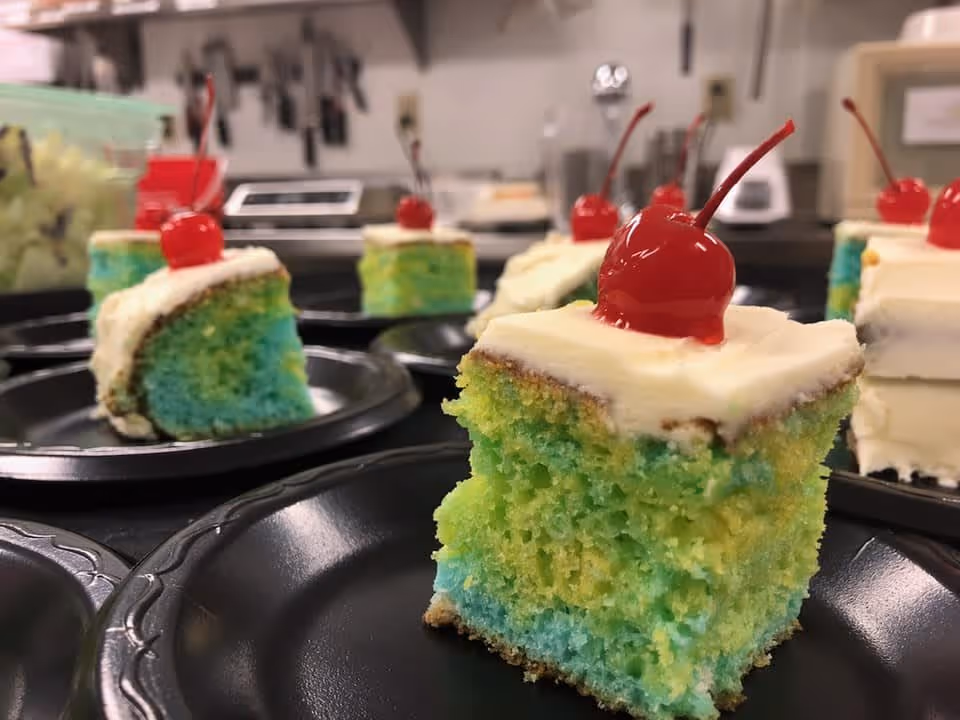 Colorful green-and-blue cake squares topped with white frosting and maraschino cherries on black plates in a kitchen prep area.
