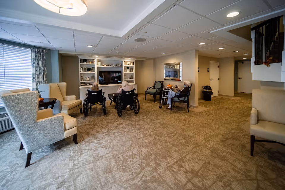 A cozy common area in a senior living facility with beige carpet and neutral-colored walls. Two elderly women in wheelchairs watch a television mounted on a white built-in shelving unit filled with decorative items. Another elderly woman with a walker sits on a chair nearby, reading a book. The room is furnished with several armchairs and has a large window with patterned curtains letting in natural light.