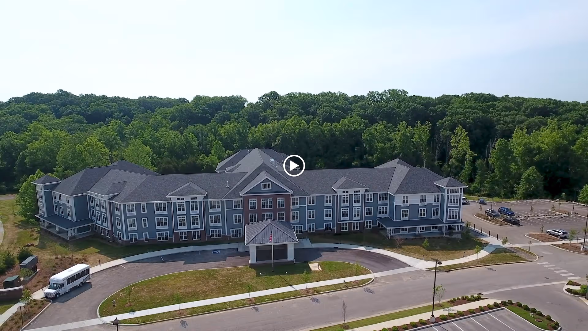 Aerial view of a large, multi-story senior living facility named Evergreen Village at Bloomington, surrounded by trees and greenery. The building has a gray exterior with white trim and a covered entrance. There is a circular driveway in front with a white shuttle bus parked on the left side and a parking lot on the right.