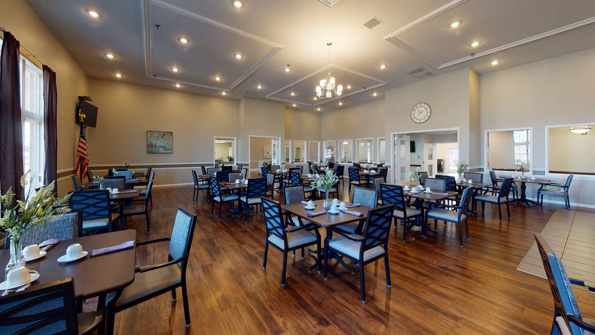 A spacious dining room in a senior living facility with multiple tables and chairs arranged neatly. Each table is set with cups, saucers, and napkins. The room has wooden flooring, beige walls, large windows with dark curtains, a chandelier, recessed ceiling lights, and a wall clock. There is an American flag in the corner and some framed artwork on the walls.