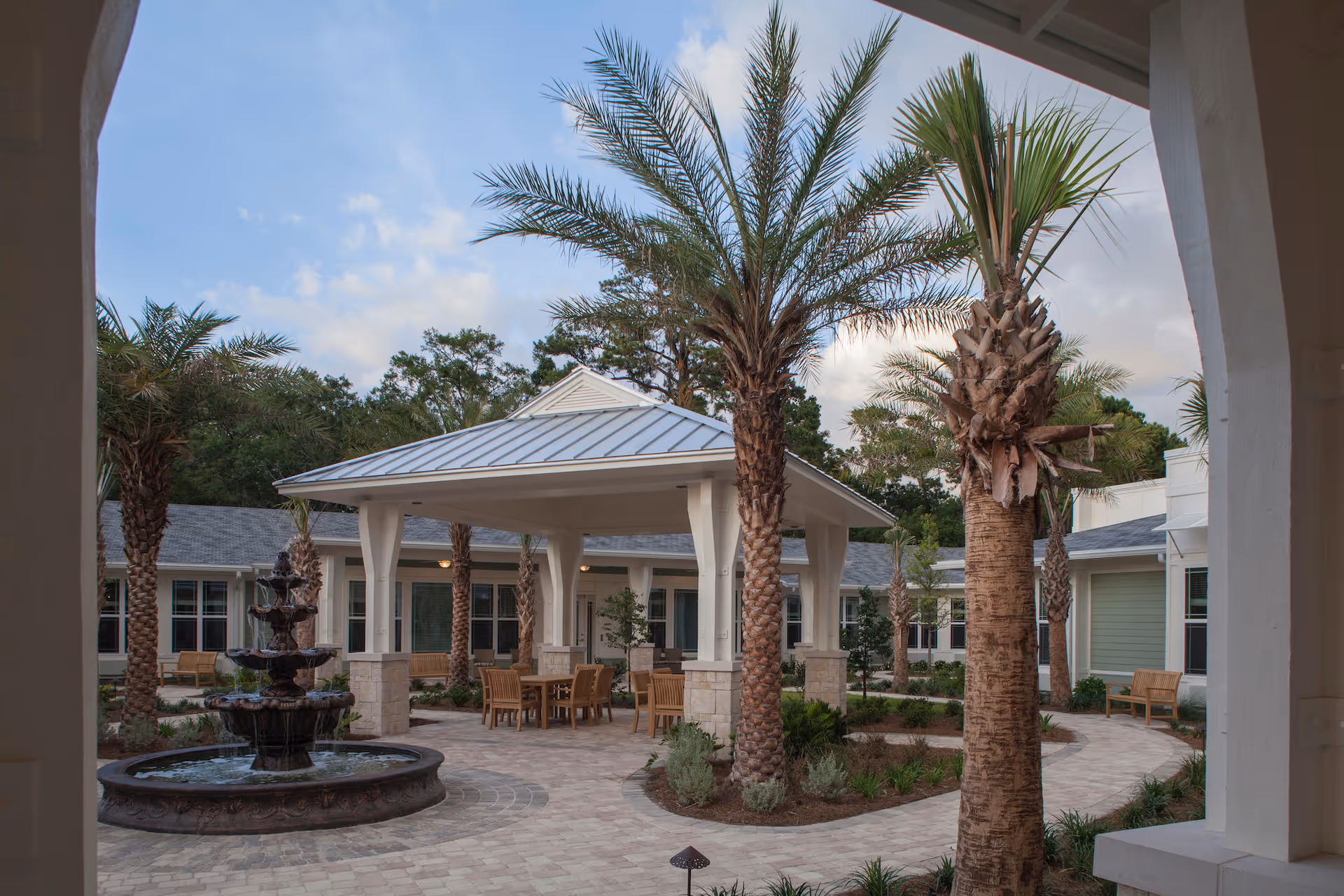 Courtyard with palm trees, a multi-tiered fountain, a covered pavilion with tables and chairs, and surrounding single-story buildings.