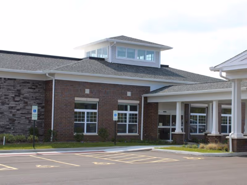 Exterior view of Avenue at North Ridgeville Care and Rehabilitation Center showing a brick building with large windows, a covered entrance supported by white columns, and a parking lot with designated handicapped parking spaces.