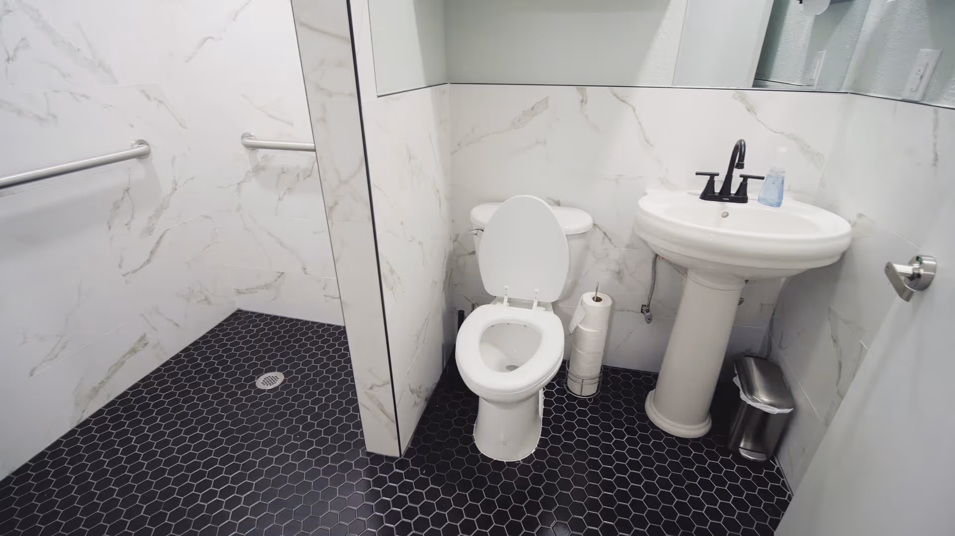 A clean bathroom with white marble-patterned walls and black hexagonal tile flooring. The bathroom features a white toilet with the lid open, a white pedestal sink with a black faucet, a soap dispenser on the sink, a metal trash can, a toilet paper holder with multiple rolls, and a stainless steel grab bar on the wall near a walk-in shower area.