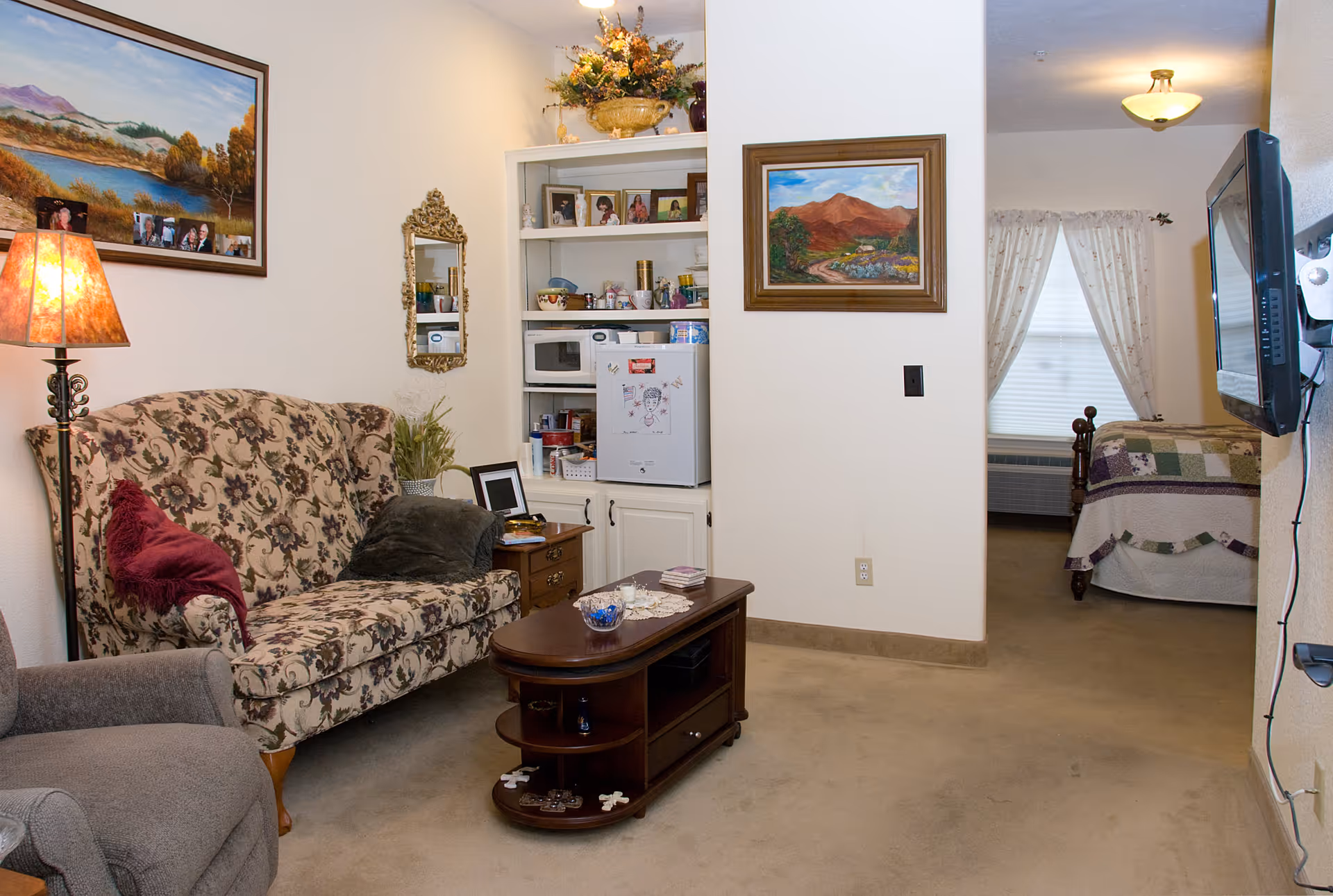 A cozy living room area with a floral patterned sofa, a gray armchair, a wooden coffee table, and a side table with a lamp. There are framed paintings and family photos on the walls and shelves. In the background, a bedroom with a bed covered in a patchwork quilt is visible through an open doorway.