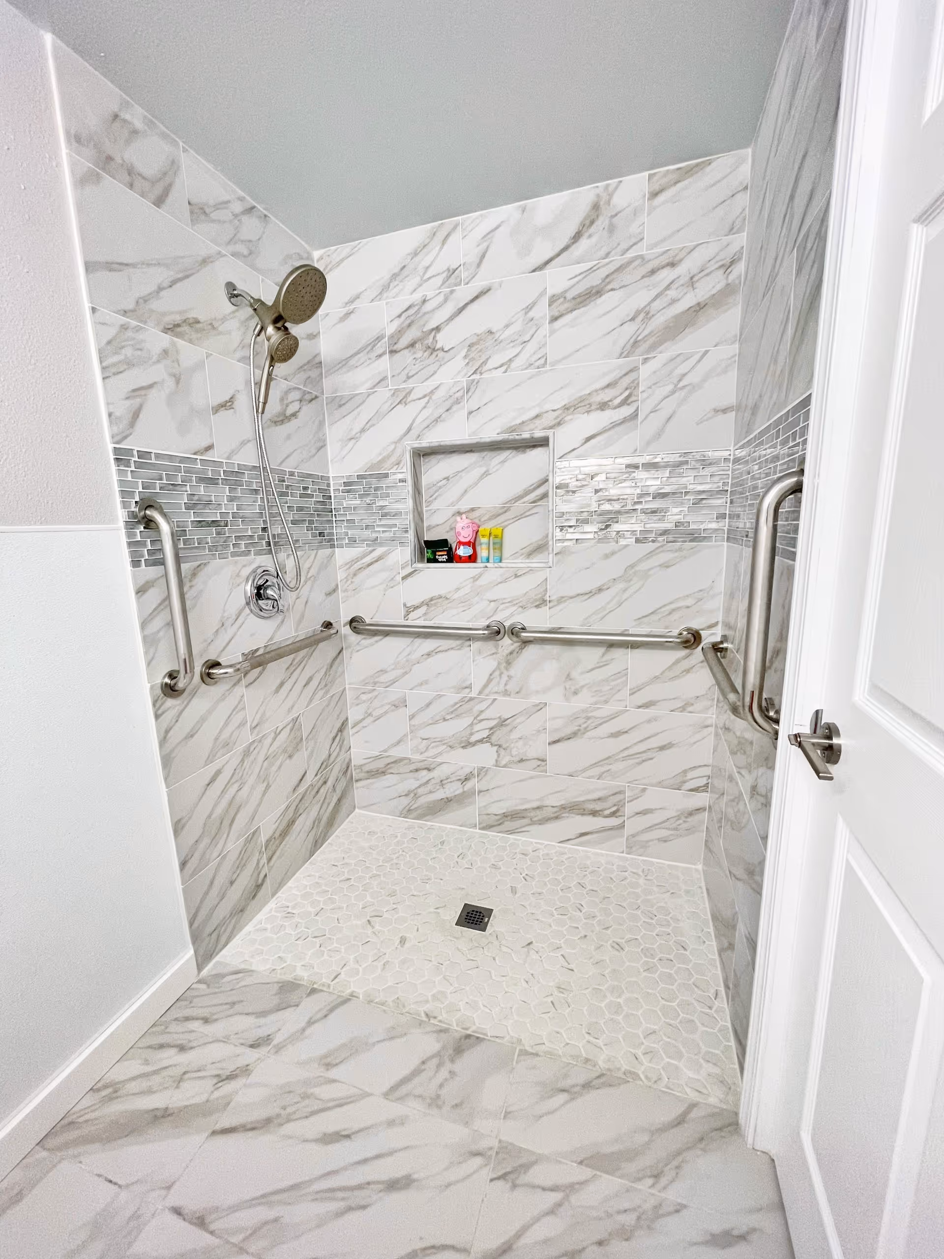 Tiled walk-in shower with marble-look tiles, grab bars, a handheld showerhead, and a built-in niche holding toiletries.