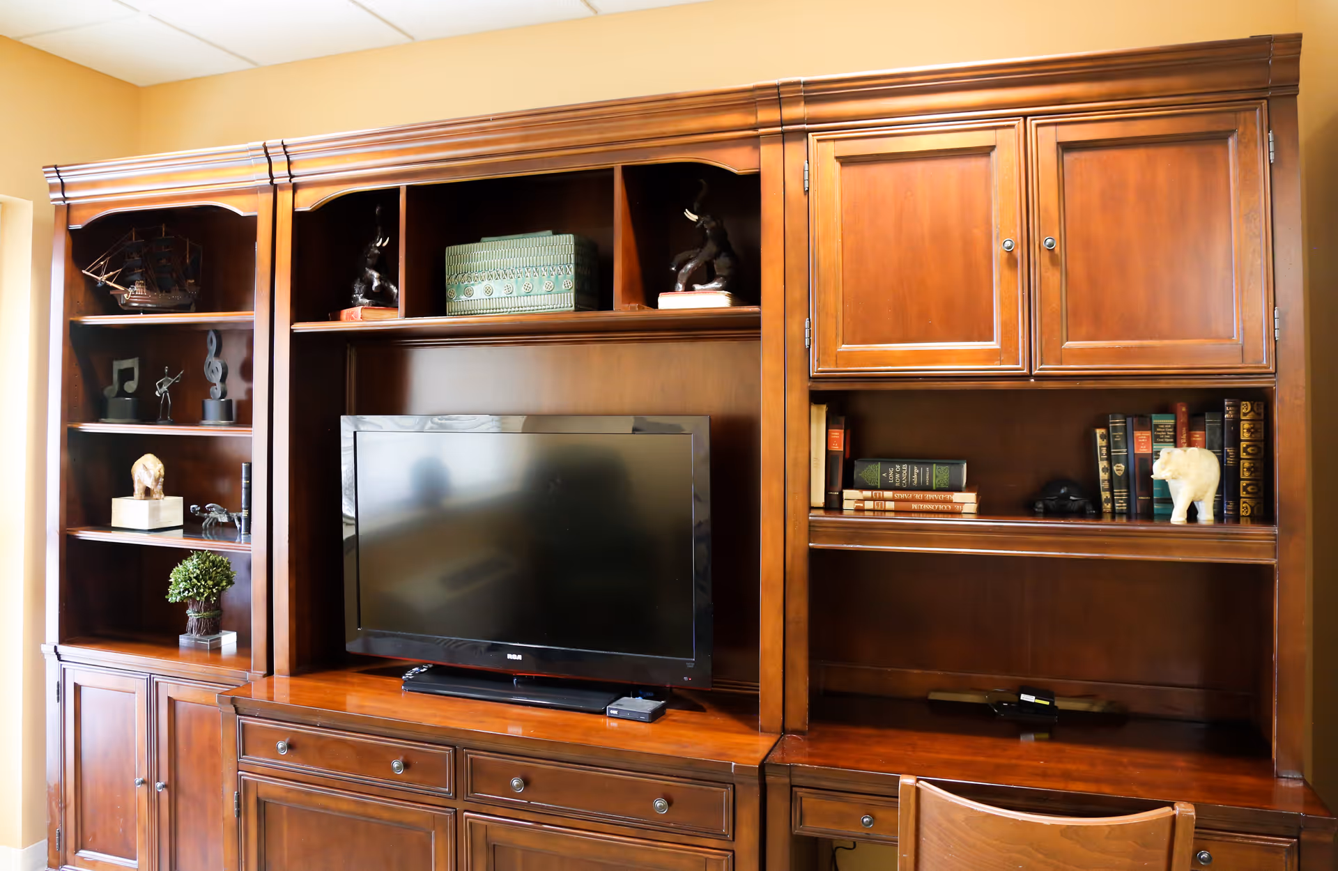 A wooden entertainment center with shelves and cabinets, featuring a flat-screen TV in the center. The shelves hold decorative items including a model ship, small sculptures, books, and a small potted plant. There is a wooden chair partially visible in front of the right side of the unit.