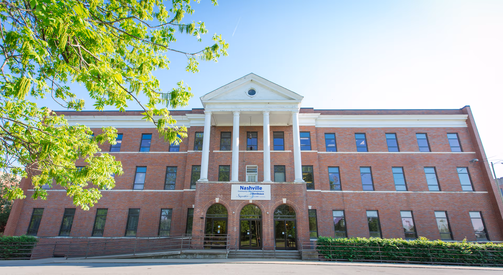 Front exterior view of a three-story red brick building with white columns and a triangular pediment above the entrance. The building has many windows and a sign that reads 'Nashville Community Care & Rehabilitation at Bordeaux'. Green trees and bushes are visible around the building under a clear blue sky.