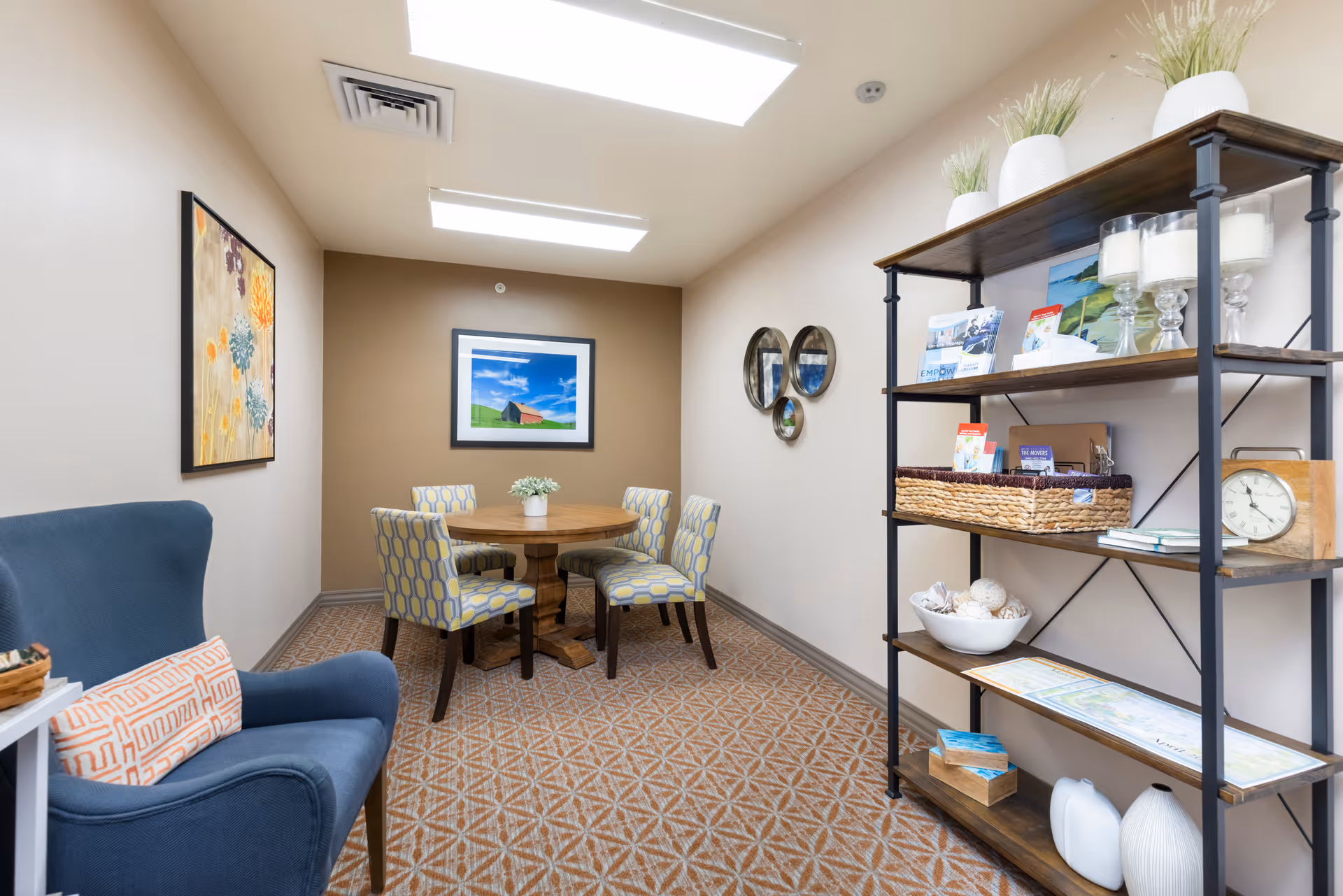 Small seating area with a round wooden table and four patterned chairs, a blue armchair, and a shelving unit in a warmly lit interior.