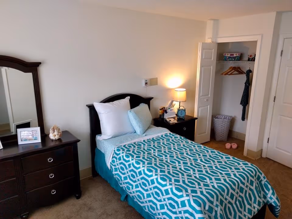 A neatly made single bed with a turquoise and white geometric patterned bedspread and multiple pillows in a bedroom. Next to the bed is a dark wooden nightstand with a lamp, alarm clock, and some small items. To the left is a matching dark wooden dresser with a mirror, a framed photo, and a decorative item. An open closet with hangers, a box, a laundry basket, and a pair of pink slippers is visible on the right side of the room.