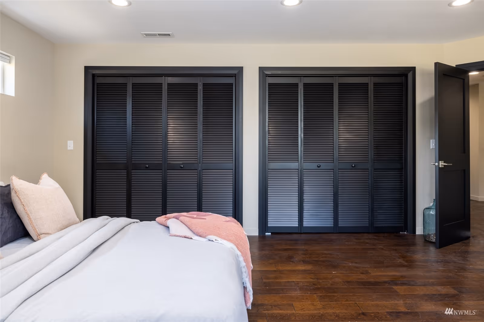 A bedroom with a bed on the left side covered with light gray and pink bedding. The room features two large black louvered closet doors on the far wall and a black door on the right side leading to another room. The floor is dark wood, and the walls are painted light beige.