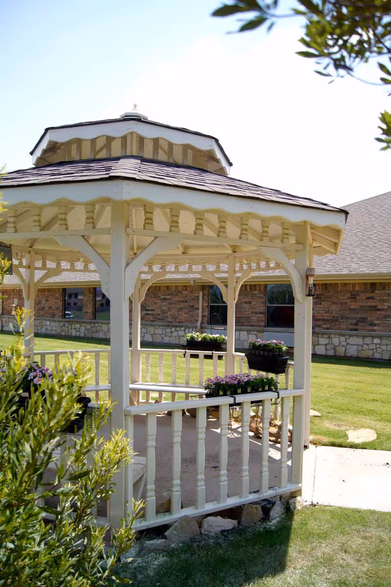A white wooden gazebo with a shingled roof situated on a grassy lawn. The gazebo has flower boxes with pink and purple flowers attached to the railing. In the background, there is a brick building with stone accents and windows. Some green shrubs are visible in the foreground.