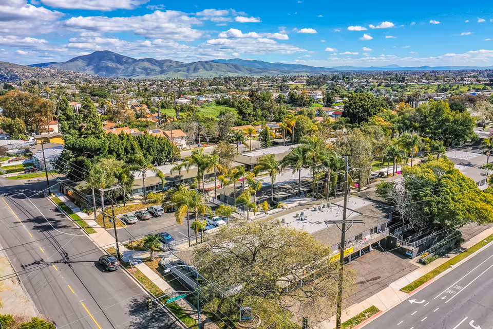 Aerial view of Monte Vista Village facility surrounded by palm trees and other greenery, with a parking lot and streets visible. The background shows a sprawling suburban area with hills and mountains under a partly cloudy sky.