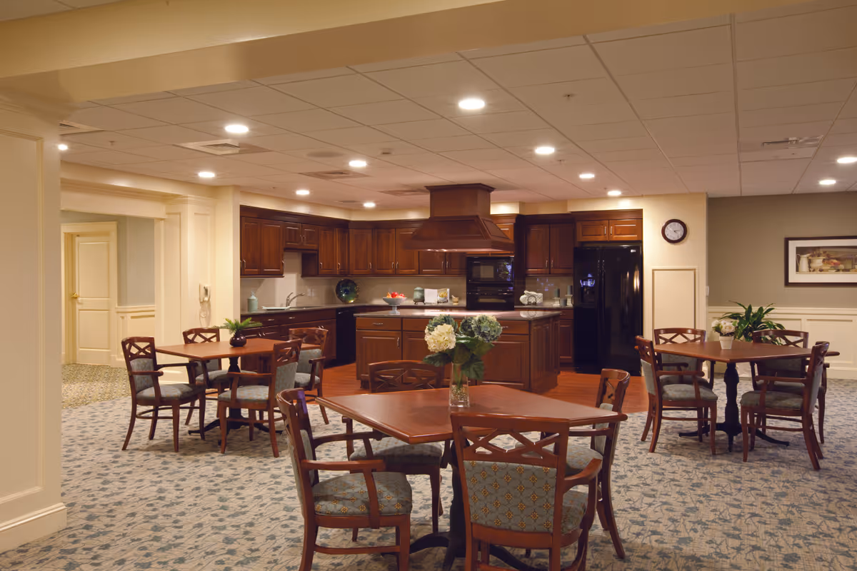 A spacious kitchen and dining area in a senior living facility featuring dark wood cabinetry, a central island with a range hood, and multiple wooden tables with cushioned chairs. The room is well-lit with recessed ceiling lights and decorated with plants and framed artwork on the walls.