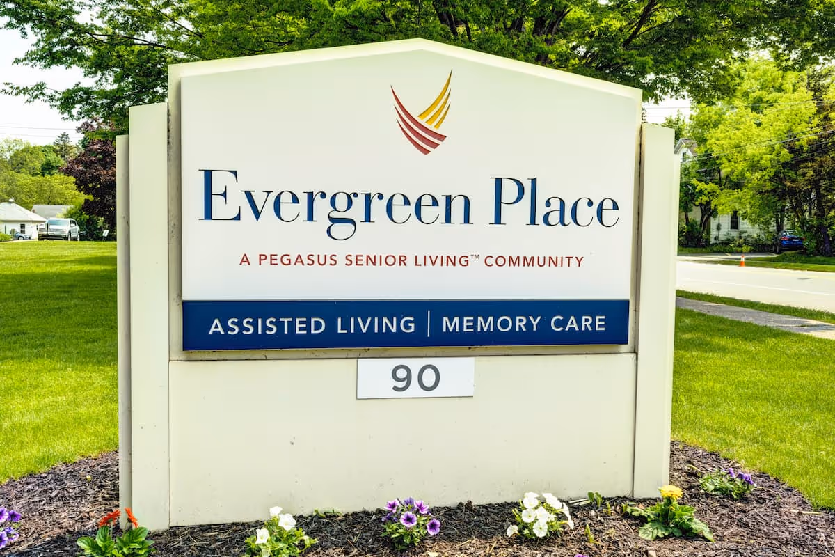 Outdoor sign for Evergreen Place, a Pegasus Senior Living community offering assisted living and memory care services, surrounded by grass and flowers with trees and a road in the background.