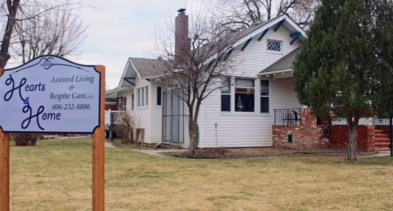 Exterior view of a white single-story house with a brick porch and a leafless tree in the front yard. A sign in the foreground reads 'Hearts & Home Assisted Living & Respite Care, LLC 406-232-8886'. The lawn is brownish-green and there are other trees around the property.