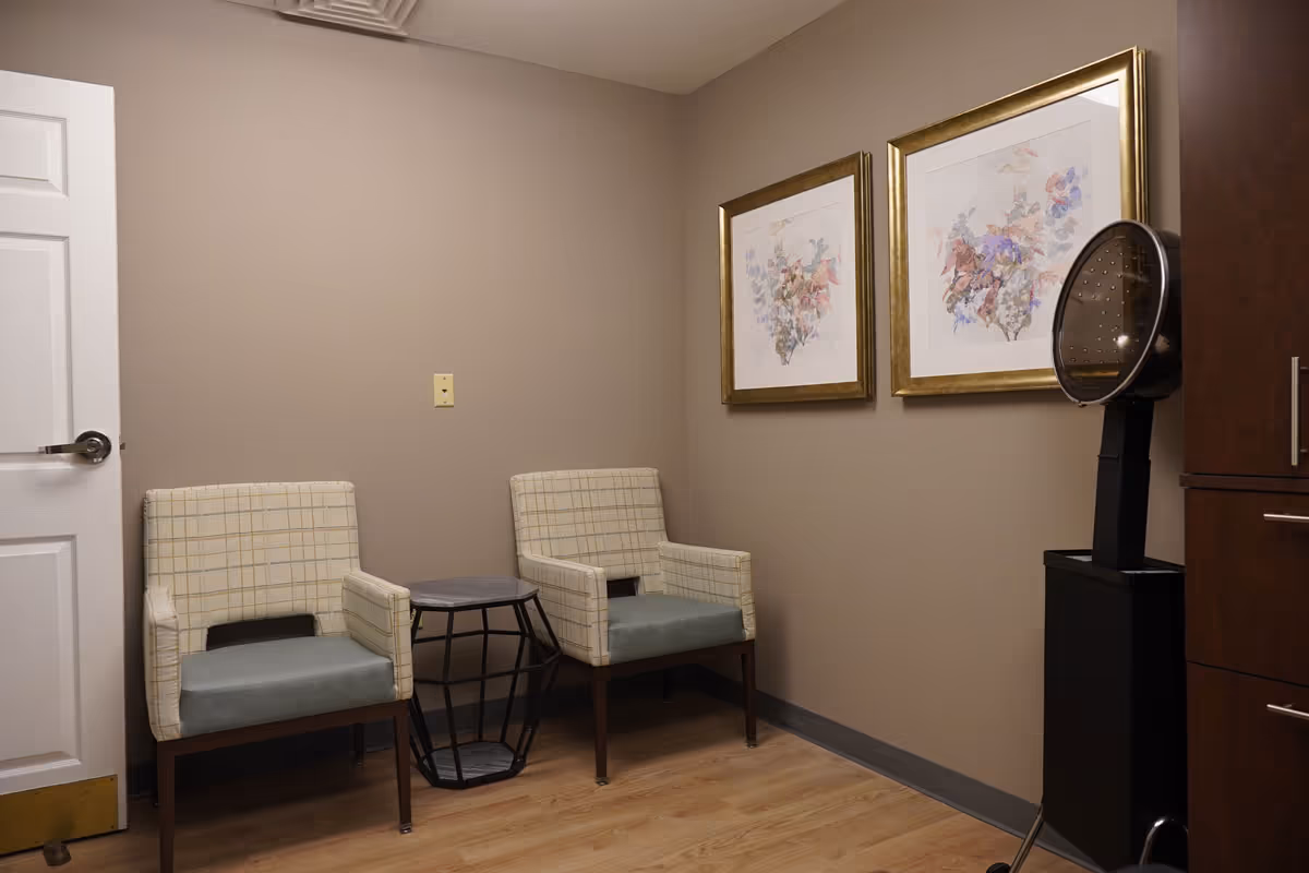 A small room with two cushioned armchairs with plaid backs and solid seats, separated by a small round side table with a geometric metal base. On the wall above the chairs are two framed floral watercolor paintings with gold frames. To the right is a black hair dryer hood on a stand next to a wooden cabinet with drawers. The walls are painted beige and the floor has light wood laminate.