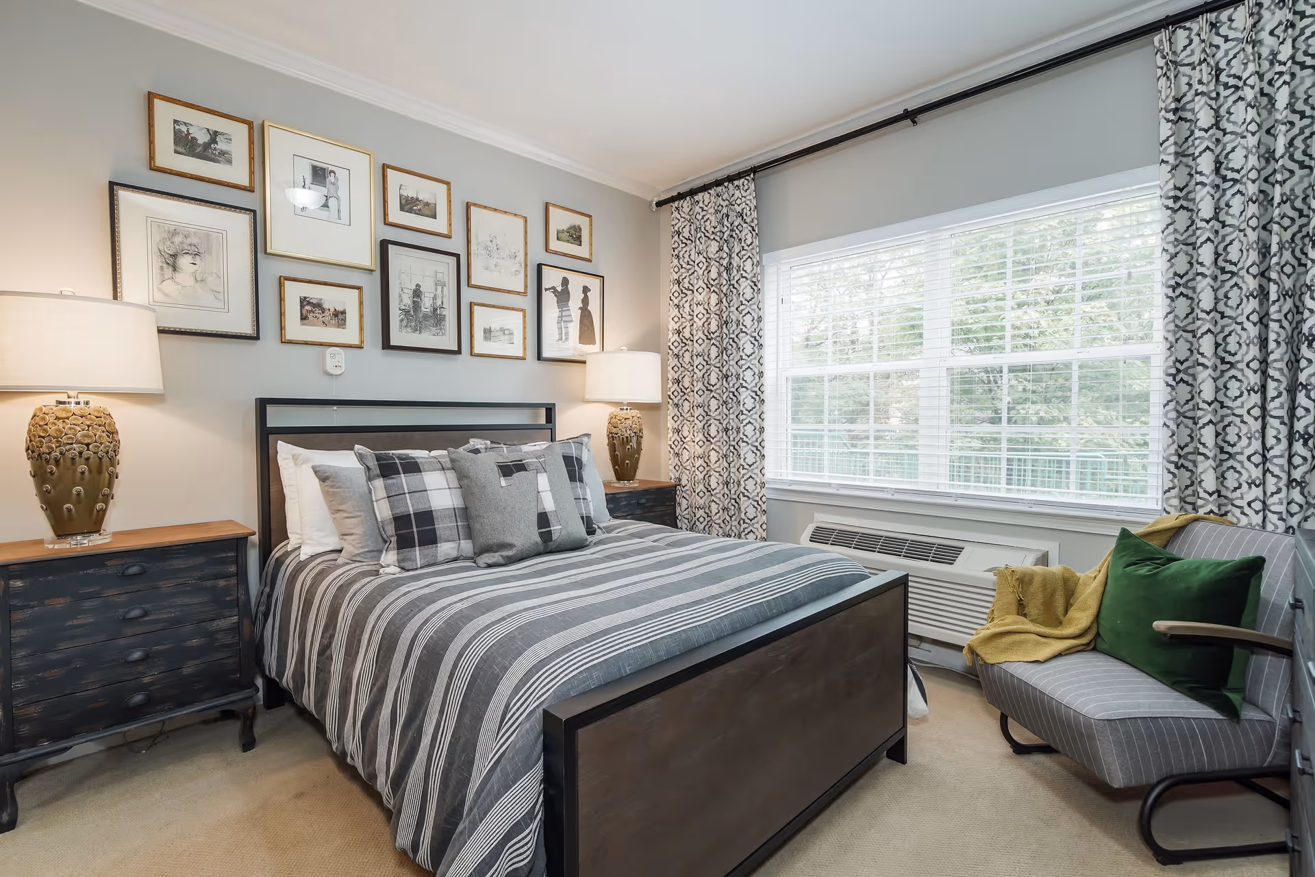 A cozy bedroom with a bed featuring striped bedding and multiple pillows. Two matching bedside tables with lamps flank the bed. Above the bed is a gallery wall with various framed artworks. A large window with patterned curtains lets in natural light. A gray armchair with a green pillow and yellow throw blanket is positioned near the window.