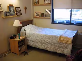 A small bedroom with a bed, nightstand, lamp, and wall photos near a window.