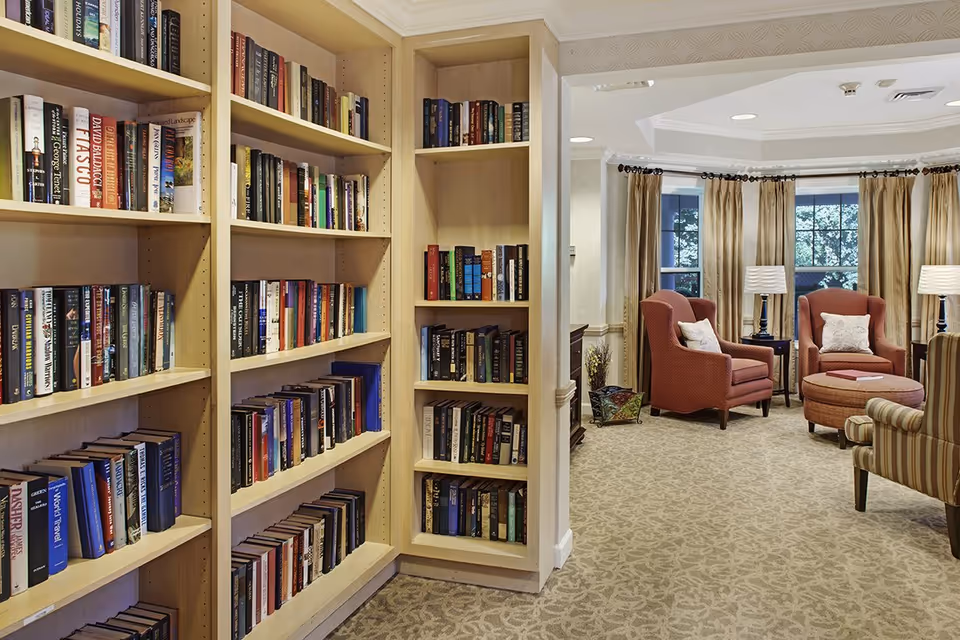 Interior view of a senior living facility library area with wooden bookshelves filled with books on the left and a cozy sitting area with armchairs, ottoman, side tables, lamps, and large windows with curtains on the right.