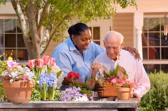 A caregiver and an elderly man are gardening together outdoors, tending to various colorful potted flowers on a wooden table. The caregiver is smiling and gently guiding the elderly man, who is also smiling. They are in a garden area with a tree and a building with windows in the background.