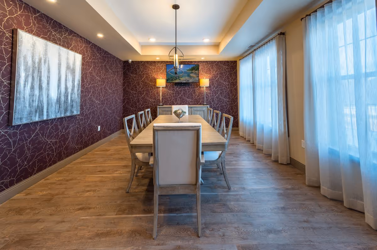 A dining room with a long wooden table surrounded by eight chairs. The room has wooden flooring, a purple accent wall with a large abstract painting, and two lamps on a sideboard beneath a mounted TV. Large windows with sheer curtains allow natural light to fill the space.