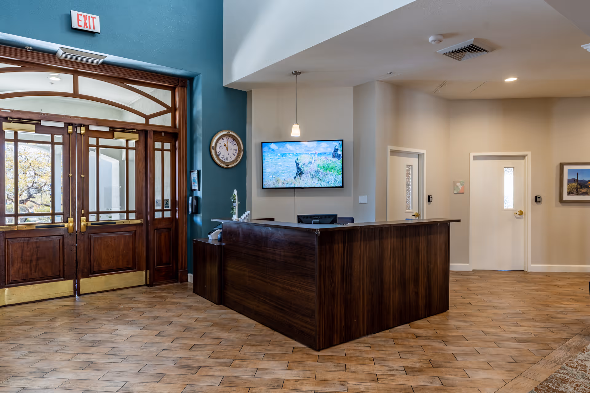 Reception area with a dark wooden front desk, a wall-mounted TV displaying a nature scene, a round wall clock, and double wooden doors with glass panels. The floor is tiled with a wood-like pattern, and there are two white doors and a framed picture on the beige walls.