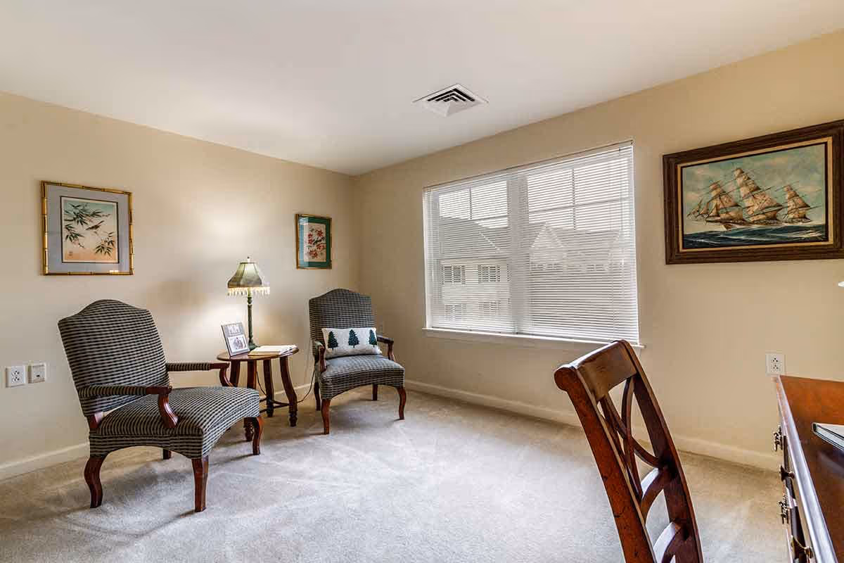 Sunny seating area with two striped armchairs, a round side table with a lamp, a window with blinds, and framed artwork on the walls.