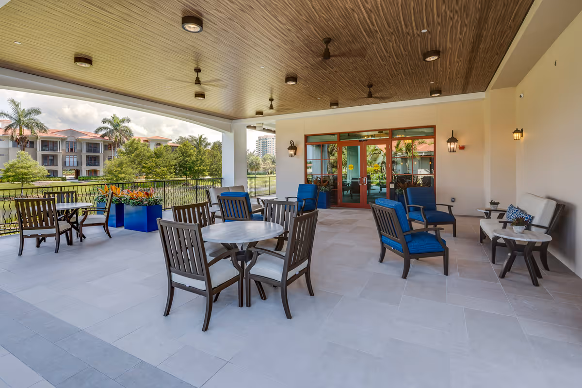Covered outdoor patio area with several seating arrangements including wooden chairs with cushions around round tables and blue cushioned armchairs. The patio overlooks a landscaped area with trees and buildings in the background. The ceiling has recessed lighting and ceiling fans, and there are wall-mounted lantern-style lights near glass doors leading inside.