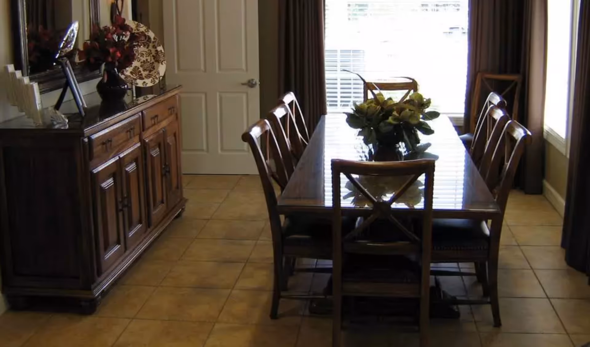 A dining room with a wooden dining table and six matching chairs. The table has a glass top and a floral centerpiece. There is a wooden sideboard with decorative items including a vase with flowers and a decorative plate. The room has tiled flooring and large windows with brown curtains allowing natural light to enter.